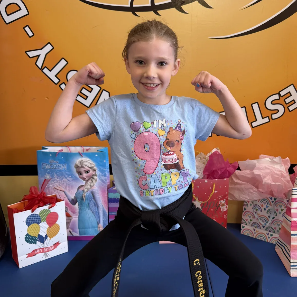 Birthday girl in her Mastery uniform flexing beside her presents at a Mastery Martial Arts birthday party